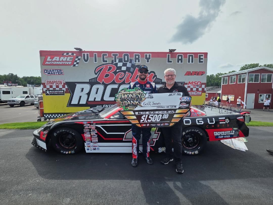 Two men standing in front of a race car and a Victory Lane Racing truck at a racetrack, holding a large check for $1,500.00.